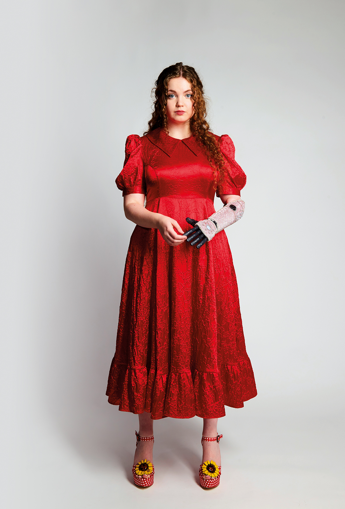 An image of Gracie McGonigal. She has long curly ginger hair and is wearing a red floral dress and red polka dot high heels with a sunflower design. She is using a bionic arm on her left which is decorated with pink sequins.
