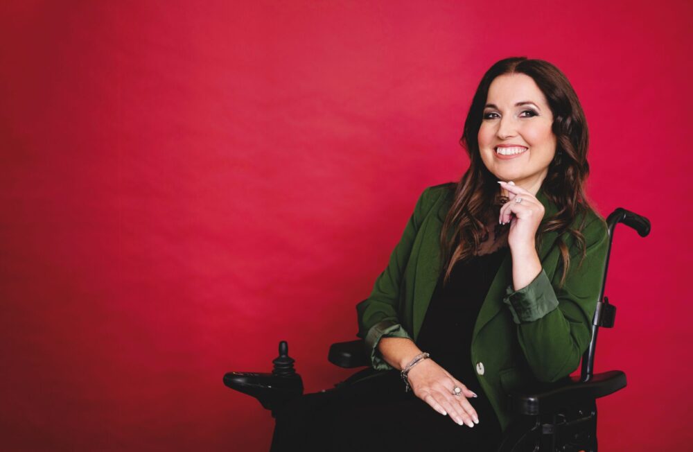 A professional image of Kim Tserkezie. She is smiling, has long brown hair and is wearing a green velvet suit jacket. She is using a wheelchair.