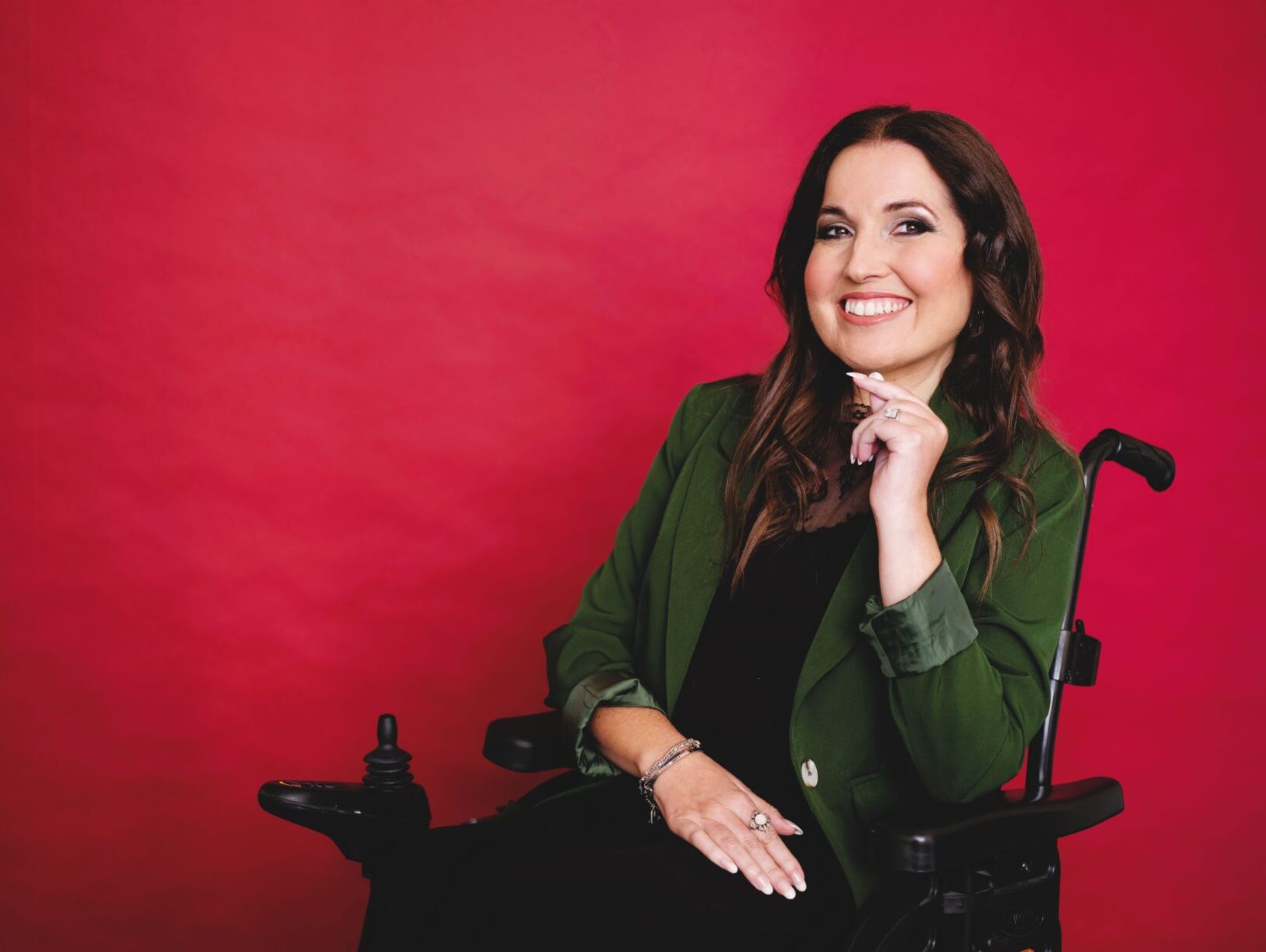 A professional image of Kim Tserkezie. She is smiling, has long brown hair and is wearing a green velvet suit jacket. She is using a wheelchair.