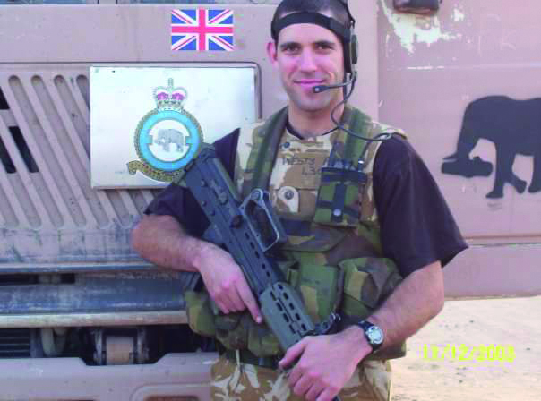 An image of Nick from his time in the Army. He is in tactical gear and is posing with a rifle in front of a lorry.