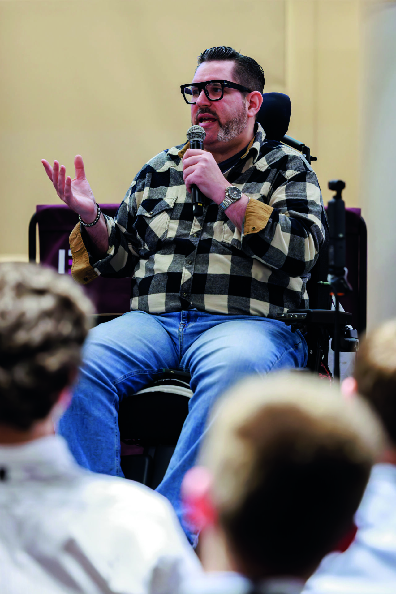An image of Nick giving a talk with a microphone to an audience. He is wearing a black white and grey plaid shirt and jeans. He is using a wheelchair.