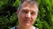 An image of Tim Rushby-Smith. He is smiling, has short grey hair, and is wearing a grey t-shirt and a wedding ring on a necklace around his neck. He is outside and behind him is a tree.