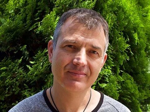 An image of Tim Rushby-Smith. He is smiling, has short grey hair, and is wearing a grey t-shirt and a wedding ring on a necklace around his neck. He is outside and behind him is a tree.