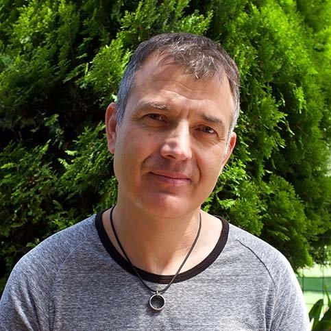 An image of Tim Rushby-Smith. He is smiling, has short grey hair, and is wearing a grey t-shirt and a wedding ring on a necklace around his neck. He is outside and behind him is a tree.