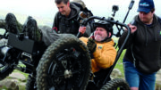 Nick Wilson is using an all-terrain powerchair to ascend a mountain. He is wearing a crash helmet, a yellow hoodie, gloves and boots. Behind him two supportive friends give the powerchair a helpful push over a tricky bit of terrain.