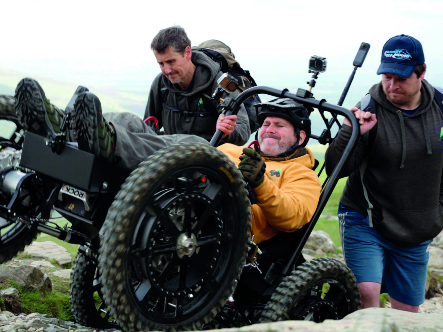 Nick Wilson is using an all-terrain powerchair to ascend a mountain. He is wearing a crash helmet, a yellow hoodie, gloves and boots. Behind him two supportive friends give the powerchair a helpful push over a tricky bit of terrain.