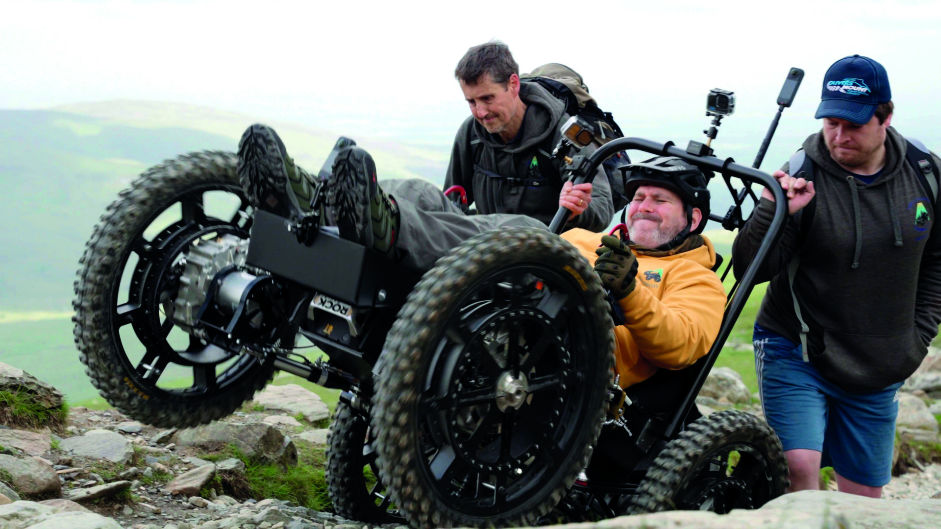 Nick Wilson is using an all-terrain powerchair to ascend a mountain. He is wearing a crash helmet, a yellow hoodie, gloves and boots. Behind him two supportive friends give the powerchair a helpful push over a tricky bit of terrain.
