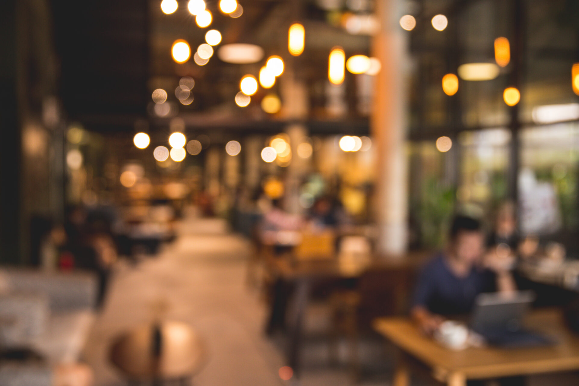 A blurred photo of a coffee shop or modern library, with an abstract bokeh light. Someone is working on their laptop with a cup of coffee.