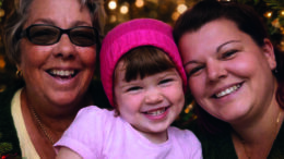 An image of Dani, her mum and her late daughter Darcy. They are all smiling and posing for a photo surrounded by Christmas trees and lights. Dani is wearing a black top, a necklace, and has long brown hair. Darcy is wearing a bright pink beanie and a pink t-shirt. Dani's mum is wearing glasses, a necklace, and a black cardigan.