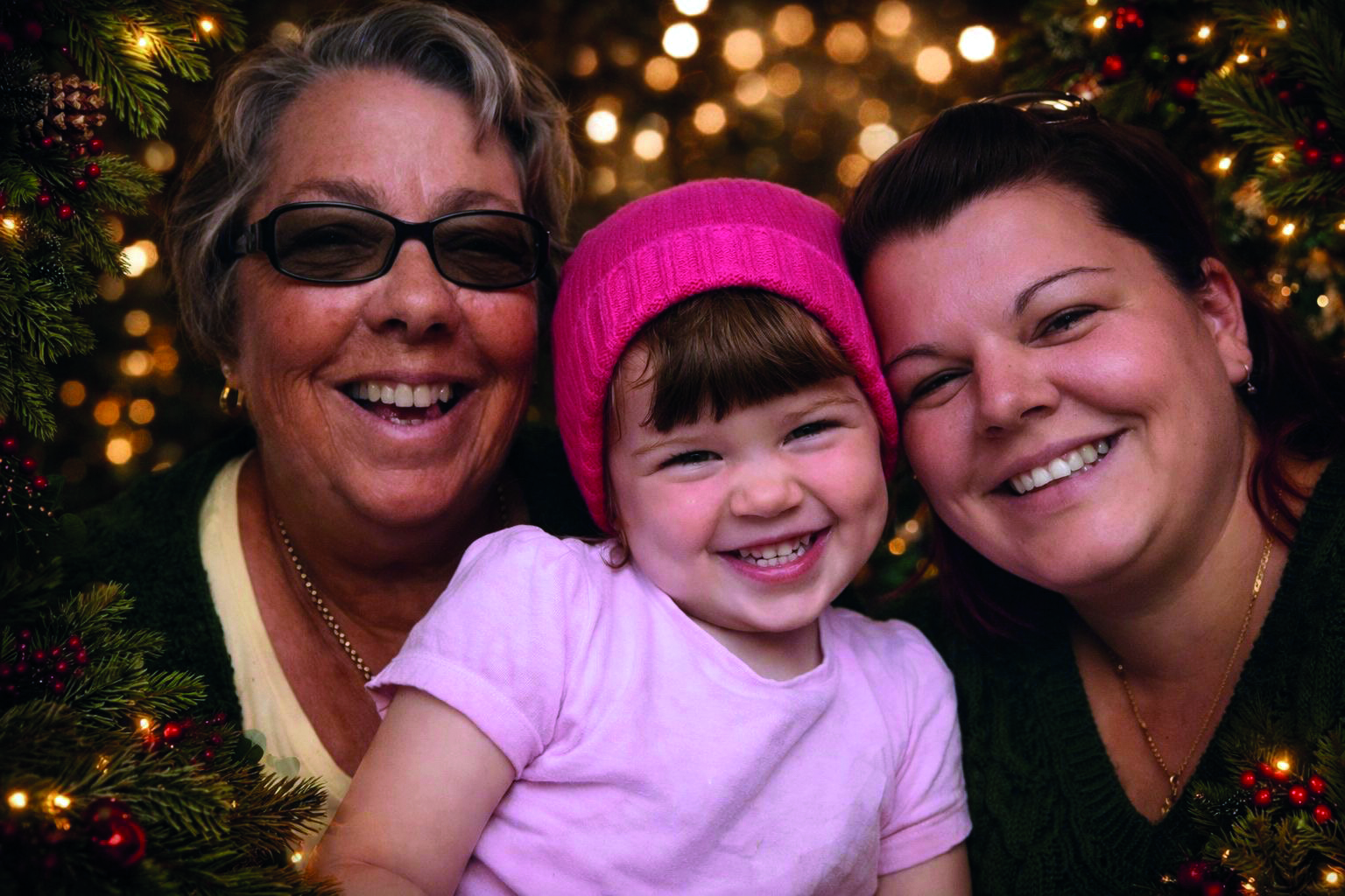 An image of Dani, her mum and her late daughter Darcy. They are all smiling and posing for a photo surrounded by Christmas trees and lights. Dani is wearing a black top, a necklace, and has long brown hair. Darcy is wearing a bright pink beanie and a pink t-shirt. Dani's mum is wearing glasses, a necklace, and a black cardigan.