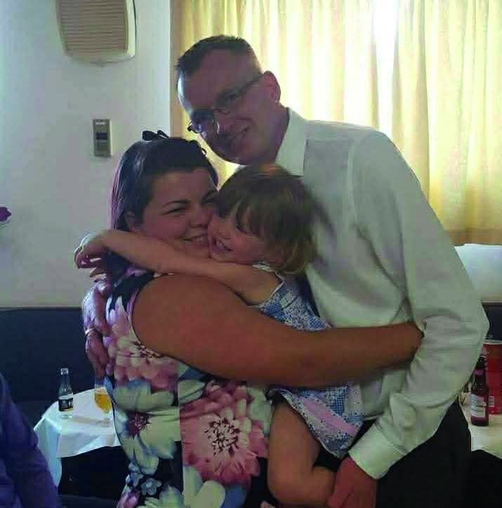 An image of Dani, Dan and Darcy. Dani is smiling and hugging Darcy who is held between her and Dan. She is wearing a pink floral dress. Darcy is smiling and wearing a blue patterned dress. Dan is smiling and wearing glasses and a white dress shirt.