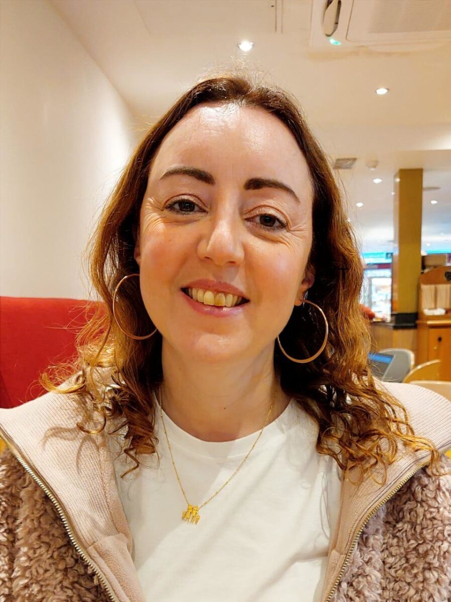 An image of Nadia Bousseau. She is smiling, has long curled brown hair and is wearing gold hoop earrings, a pink fluffy zipped hoodie and a white t-shirt.