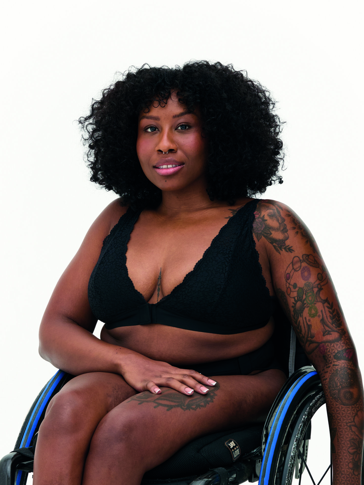 An image of a woman modelling black lingerie. She has thick black curly hair and tattoos on her left arm. She is using a wheelchair.