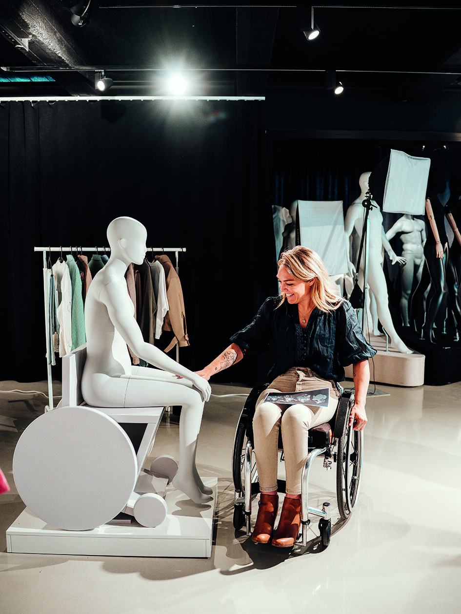 An image of a woman using a wheelchair. smiling and touching a wheelchair pose mannequin next to her, in a studio space.