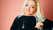 An image of Samantha Renke. She is smiling, has long blond hair and is wearing a black top and yellow floral patterned trousers. She is using a wheelchair.