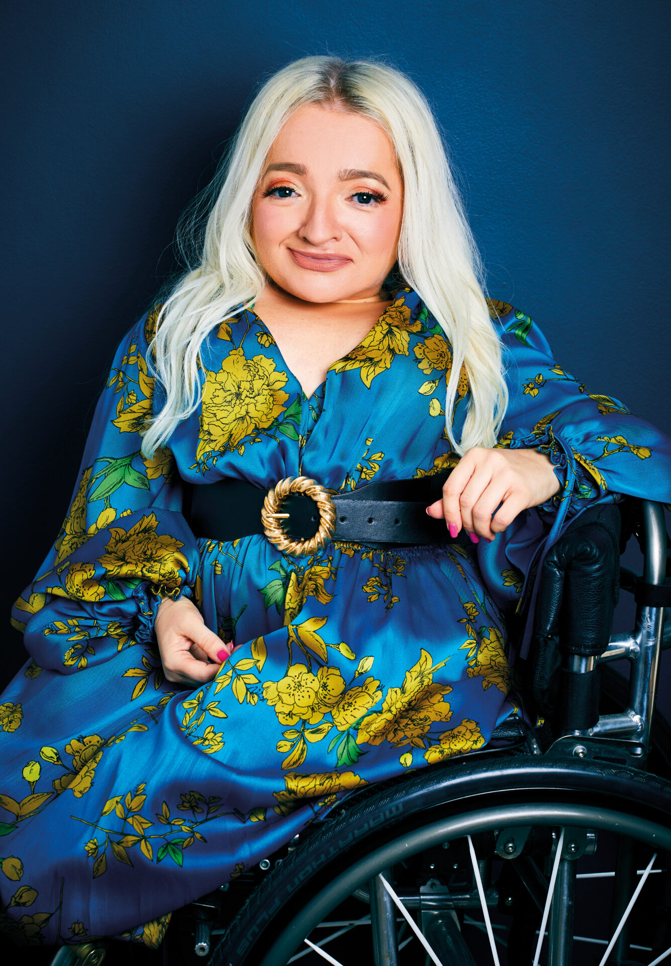 An image of Samantha Renke. She is smiling, has long blond hair and is wearing a blue and yellow floral dress. She is using a wheelchair.