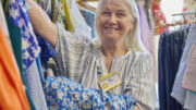 An image of a senior woman volunteering at a charity shop. She is sorting through clothes on a rail and is wearing a work badge.