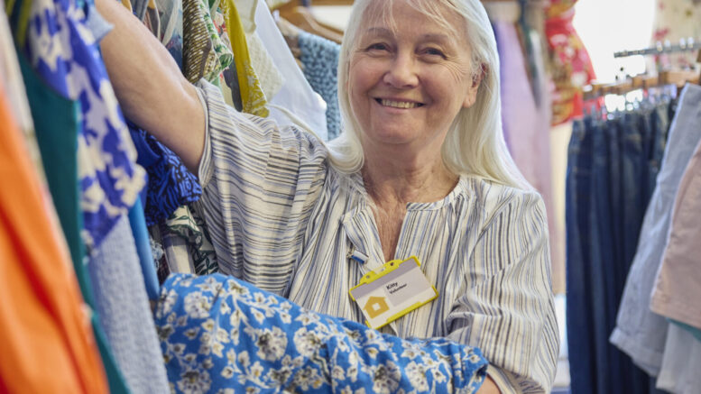 An image of a senior woman volunteering at a charity shop. She is sorting through clothes on a rail and is wearing a work badge.