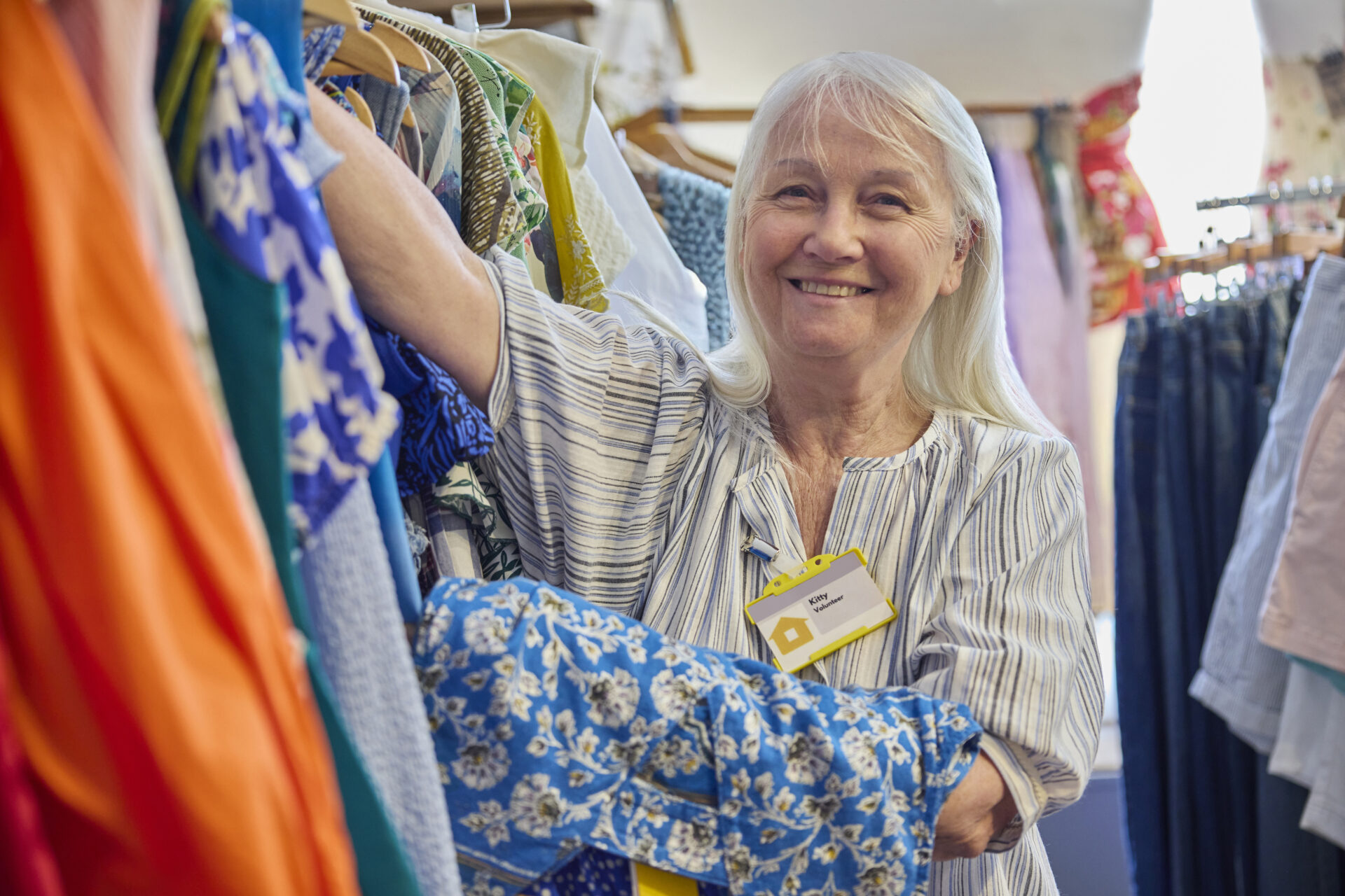 An image of a senior woman volunteering at a charity shop. She is sorting through clothes on a rail and is wearing a work badge.