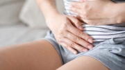 An image of a woman holding her hands over her abdomen as if in pain. She is wearing a white top with black stripes and grey shorts.