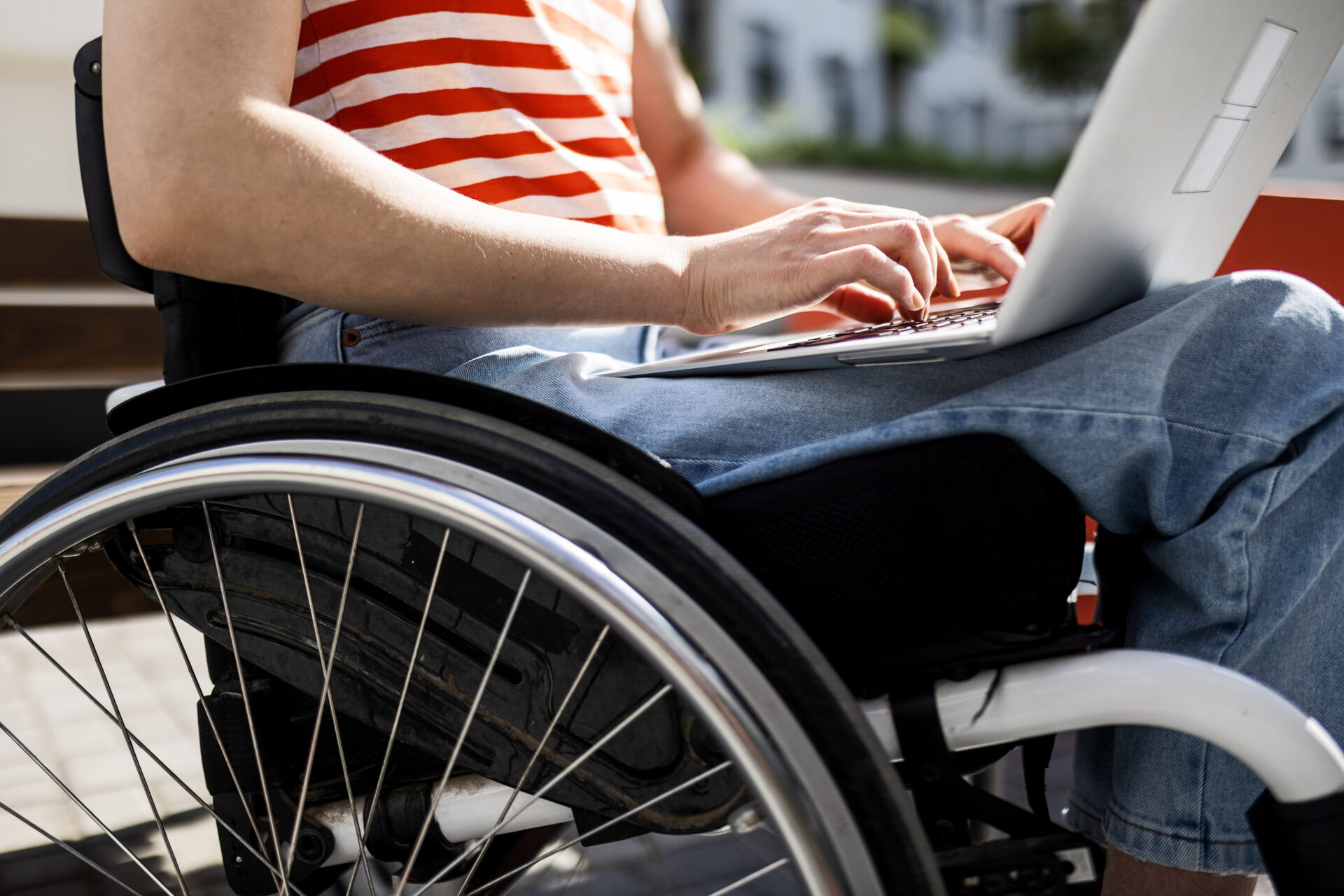 An image of an unseen woman working on a laptop. She is wearing jeans and a red and white striped t-shirt. She is using a wheelchair.