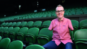 An image of Andy Stevenson. He is wearing glasses, has limb differences to his arms and legs, and is wearing a short sleeved pink shirt and formal trousers. He is wearing prosthetic legs and is sitting in the stands of a sports stadium, smiling.