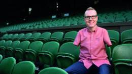 An image of Andy Stevenson. He is wearing glasses, has limb differences to his arms and legs, and is wearing a short sleeved pink shirt and formal trousers. He is wearing prosthetic legs and is sitting in the stands of a sports stadium, smiling.