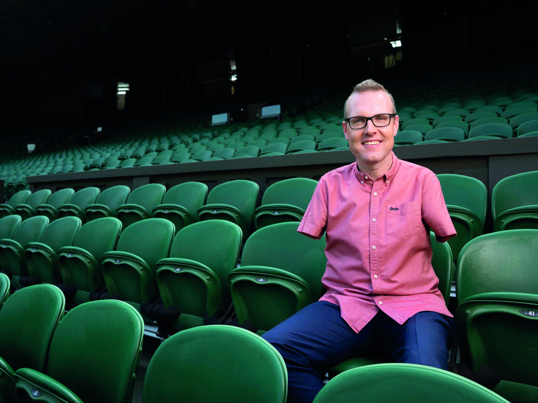 An image of Andy Stevenson. He is wearing glasses, has limb differences to his arms and legs, and is wearing a short sleeved pink shirt and formal trousers. He is wearing prosthetic legs and is sitting in the stands of a sports stadium, smiling.