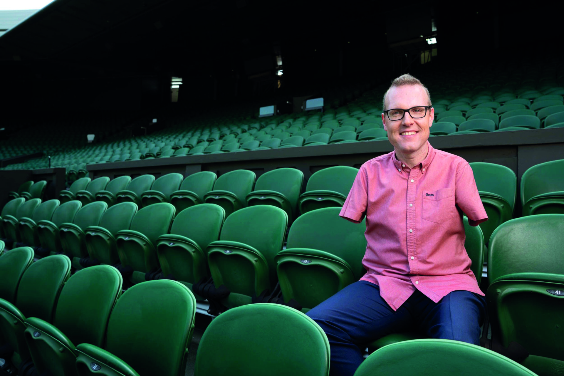 An image of Andy Stevenson. He is wearing glasses, has limb differences to his arms and legs, and is wearing a short sleeved pink shirt and formal trousers. He is wearing prosthetic legs and is sitting in the stands of a sports stadium, smiling.