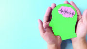 A stock image of hands holding a green human head profile cutout with a healthy brain, set against a blue background.