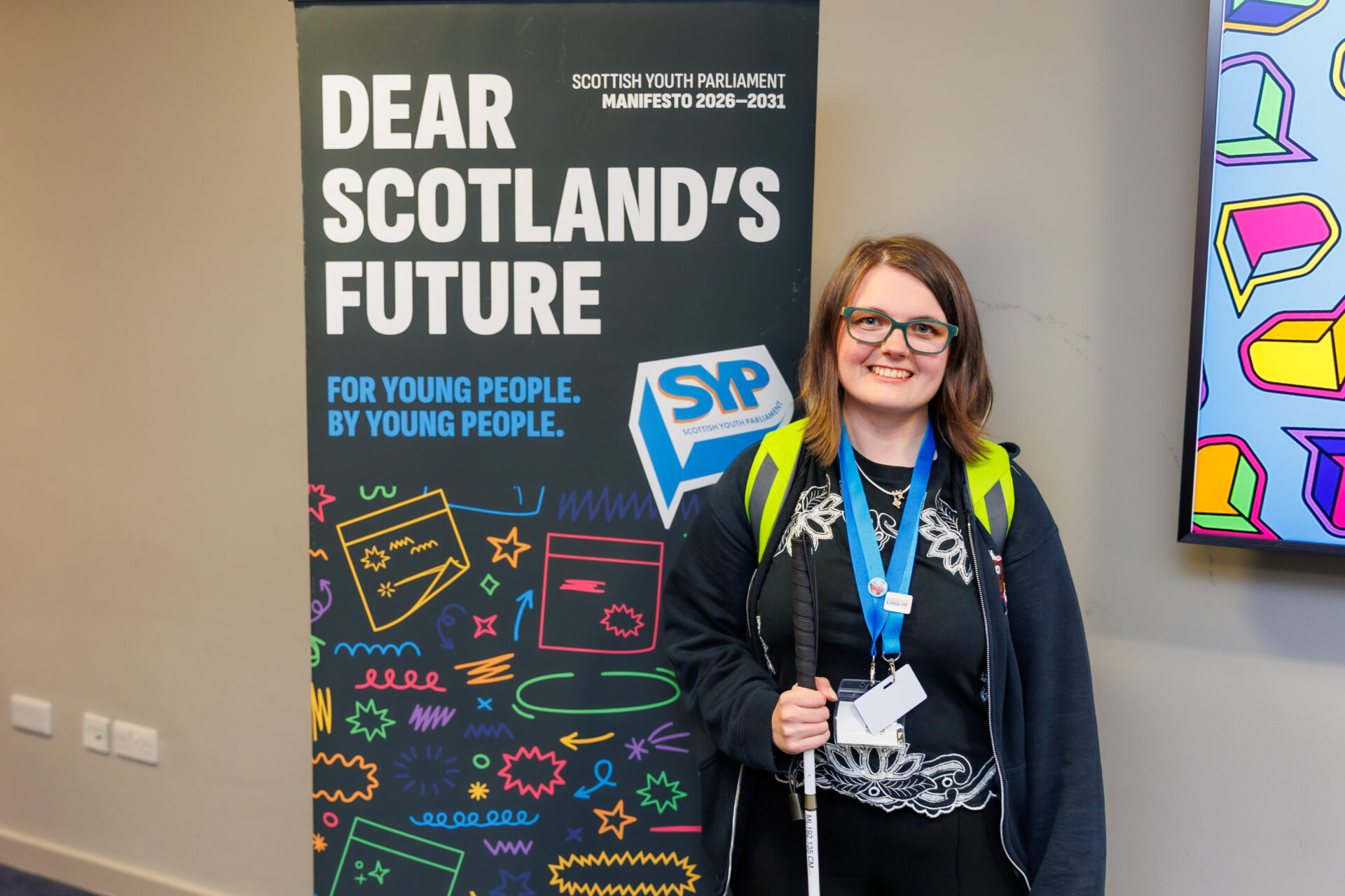 An image of Eilidh Morrison MSYP. She is smiling, has long brown hair and is wearing glasses, a black hoodie, a reflective backpack and two lanyards. She is using a cane, and standing in front of a Scottish Youth Parliament manifesto banner.
