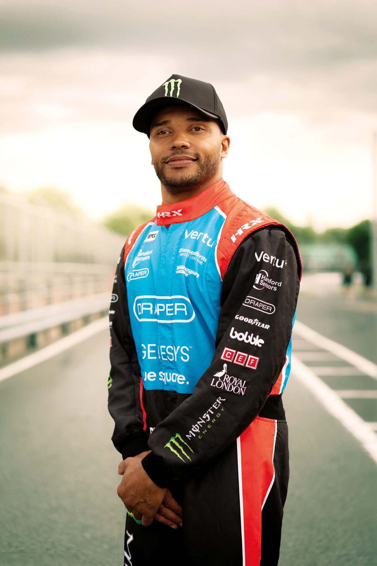 An image of Nicolas Hamilton. He is smiling and wearing a black cap and a blue, red and black racing suit. He is standing on a race track.