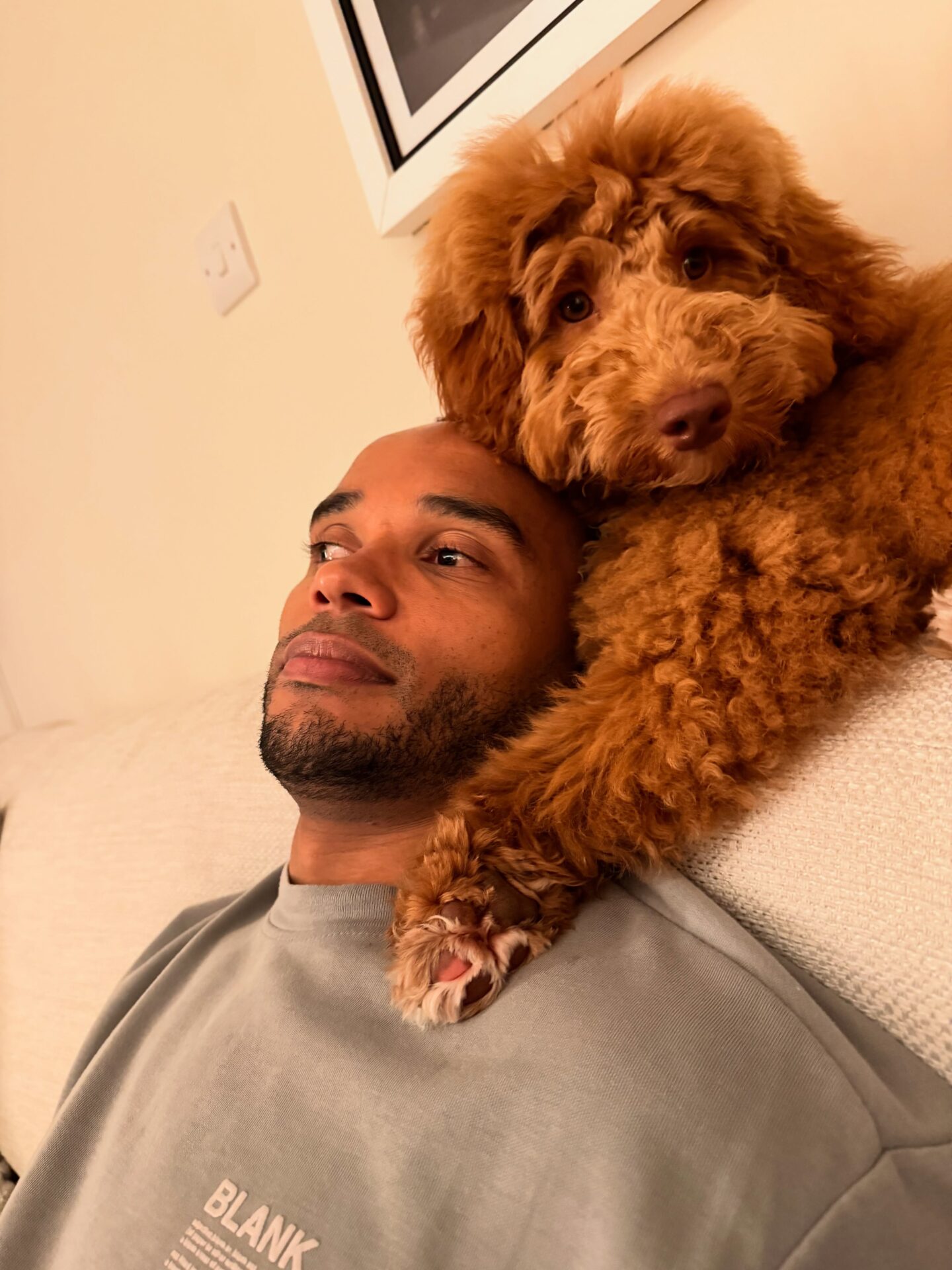 An image of Nicolas relaxing on a white couch with his dog Tippi, a red labradoodle resting her paw on his neck.