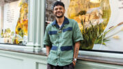 An image of Raymond Antrobus. He has black curly hair and is wearing a green and blue chequered overshirt and grey trousers.
