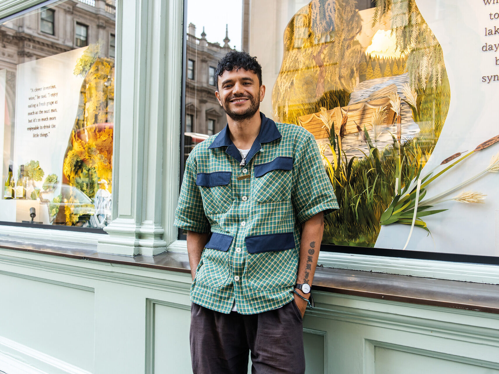 An image of Raymond Antrobus. He has black curly hair and is wearing a green and blue chequered overshirt and grey trousers.