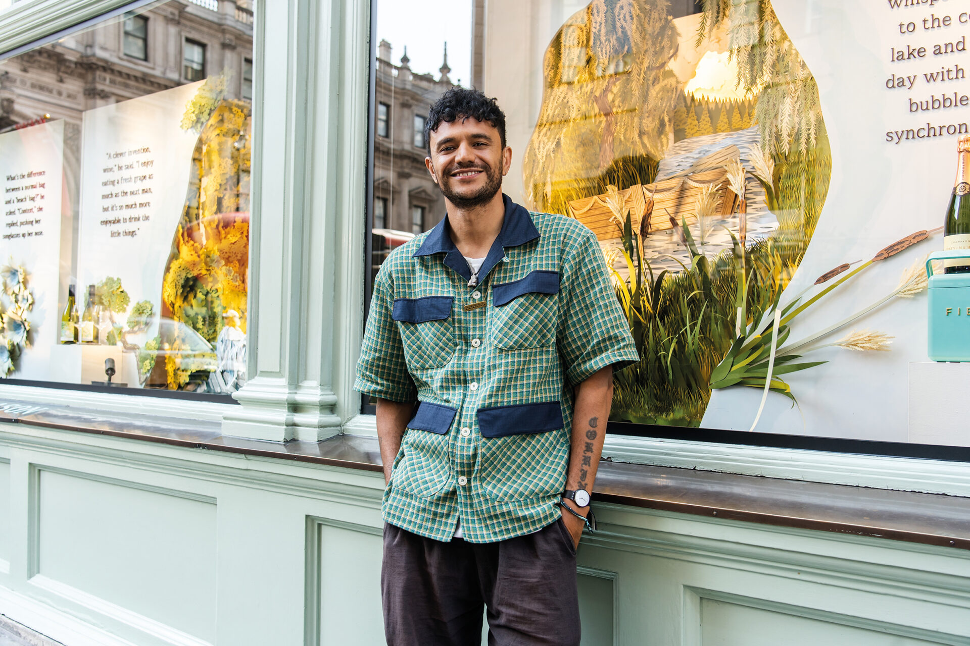 An image of Raymond Antrobus. He has black curly hair and is wearing a green and blue chequered overshirt and grey trousers. 