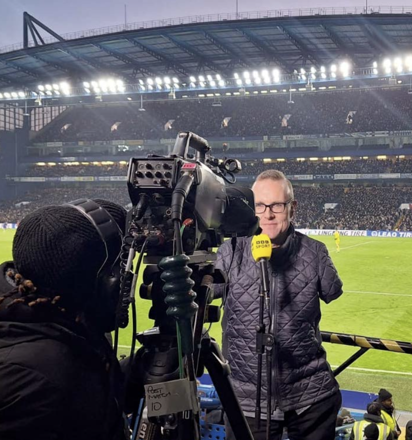 An image of Andy covering a football match, commentating in front of a camera. He is using a BBC Sport microphone and wearing a black puffer jacket.
