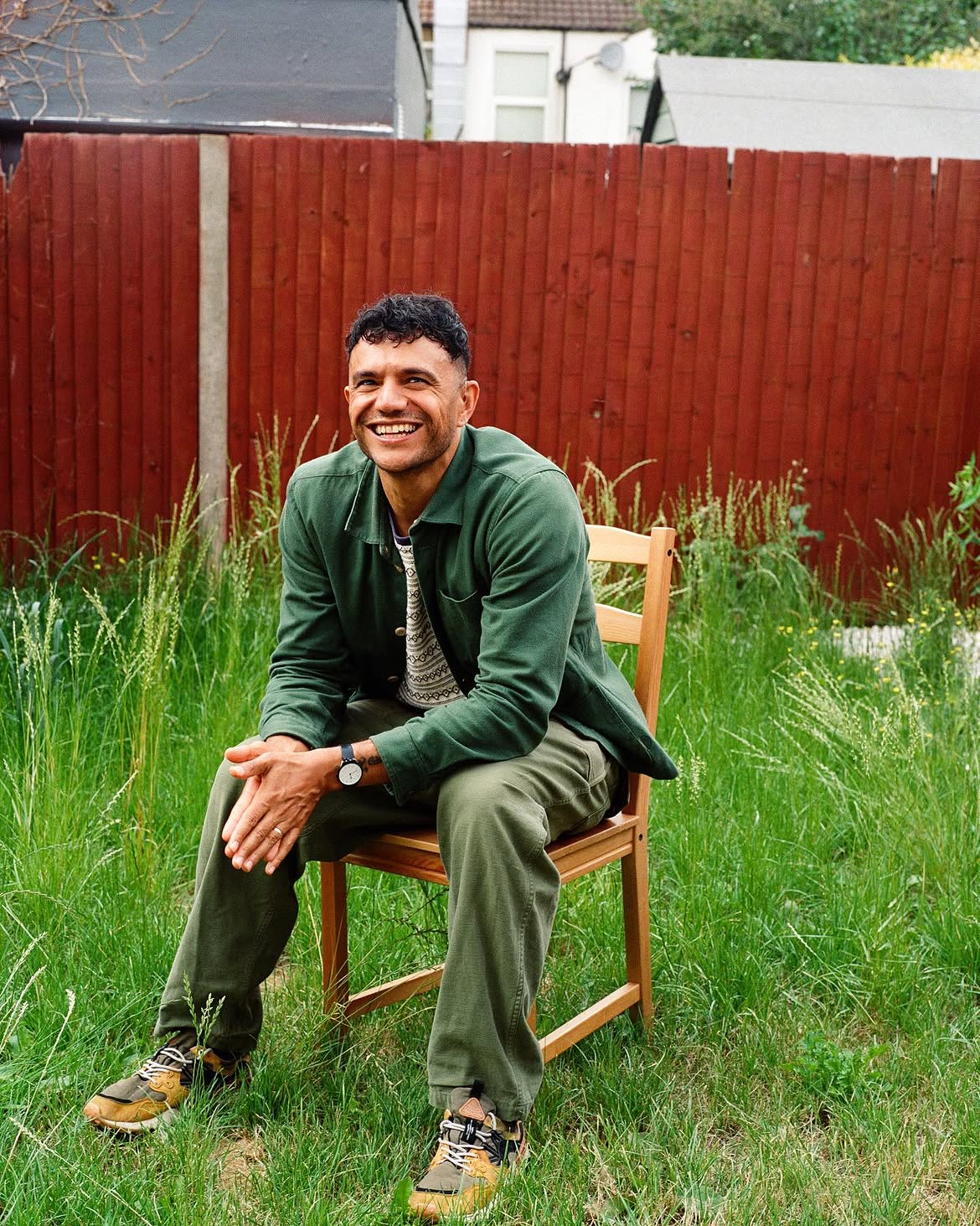 An image of Raymond smiling and sitting on a chair in a grassy garden. He is wearing a green overshirt and green jeans.