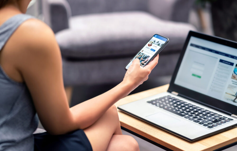 A stock image of an unknown woman looking at her phone while sitting in front of a laptop.