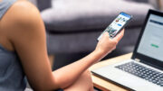 A stock image of an unknown woman looking at her phone while sitting in front of a laptop.