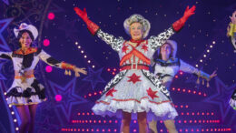 An image of Allan Stewart and company performing Jack and the Beanstalk. Allan is performing the Dame role in the panto, grinning with arms extended upwards. He is wearing a large curly grey wig, glittery patterned dress and sparkly red gloves.