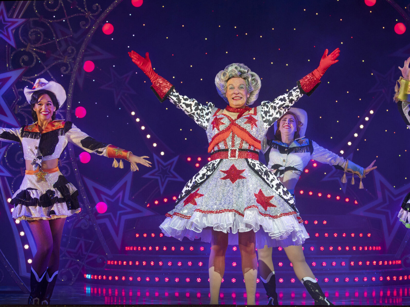 An image of Allan Stewart and company performing Jack and the Beanstalk. Allan is performing the Dame role in the panto, grinning with arms extended upwards. He is wearing a large curly grey wig, glittery patterned dress and sparkly red gloves.