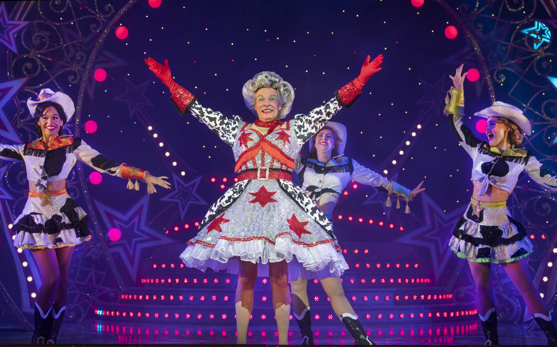 An image of Allan Stewart and other and company performing Jack and the Beanstalk. Allan is performing the Dame role in the panto, grinning with arms extended upwards. He is wearing a large curly grey wig, glittery patterned dress and sparkly red gloves.