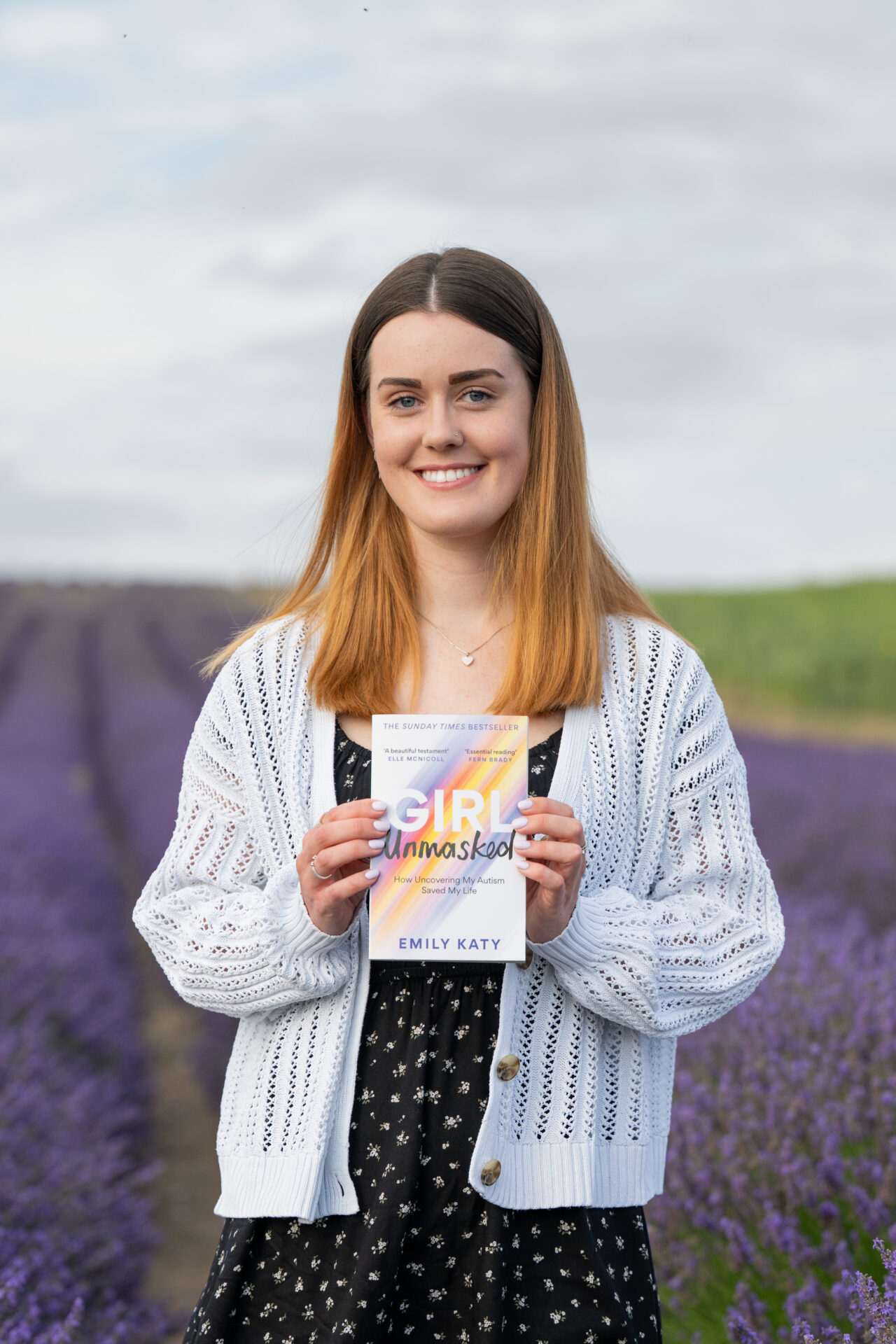 An image of Emily wearing a white cardigan and black floral dress. She is holding her book, Girl Unmasked, and standing in a field of heather.