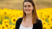 An image of Emily Katy. She is smiling, has long auburn hair and is wearing a spotted white crop top and black cardigan. She is standing in a field of sunflowers.