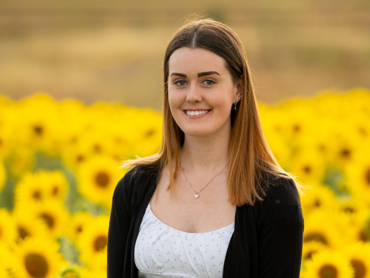 An image of Emily Katy. She is smiling, has long auburn hair and is wearing a spotted white crop top and black cardigan. She is standing in a field of sunflowers.