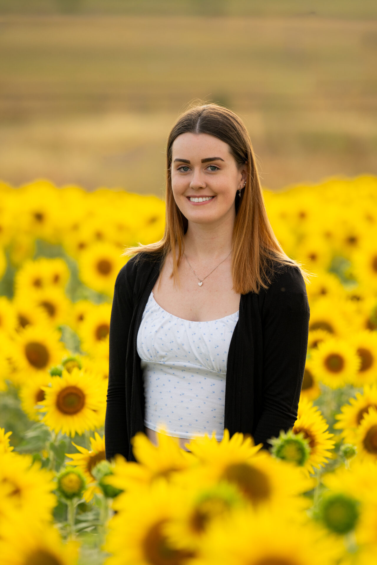 An image of Emily Katy. She is smiling, has long auburn hair and is wearing a spotted white crop top and black cardigan. She is standing in a field of sunflowers.