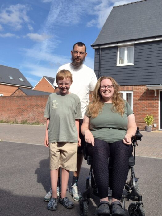 An image of Lucy and her partner and son outside a house on a sunny day. Lucy is using a wheelchair and wearing a green t-shirt and black leggings. 