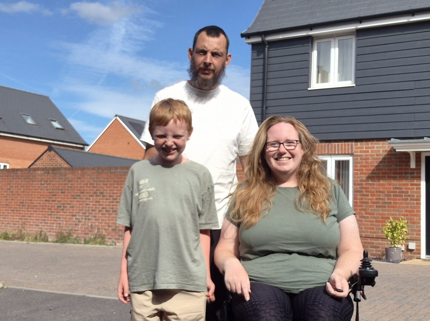 An image of Lucy Gatward and her partner and son outside a house on a sunny day. Lucy is using a wheelchair and wearing a green t-shirt and black leggings.