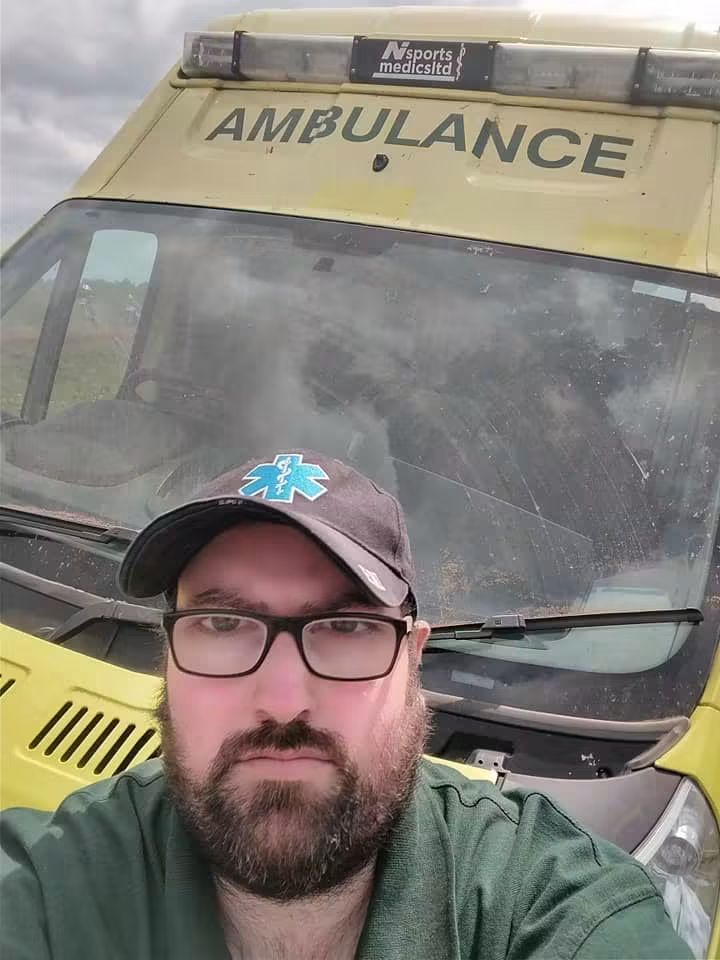 An selfie image of Martin Corey. He is wearing glasses, has a brown beard and is standing in front of an ambulance. He is wearing a paramedic's hat and uniform.